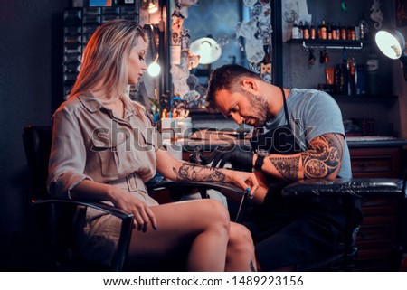 Dilligent focused tattoo artist is creating new tattoo on young woman's hand at tatoo studio.
