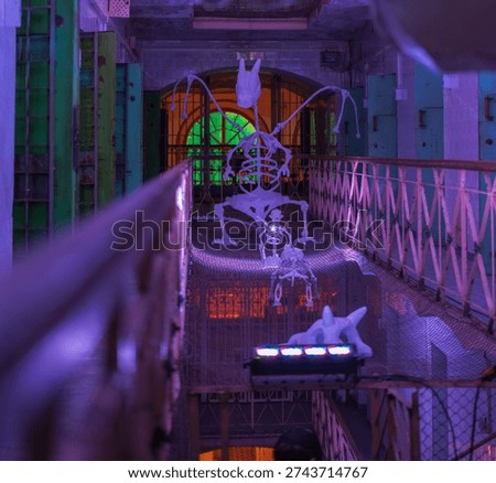 Dim corridor inside Lukiskes Prison in Vilnius, Lithuania, shows purple and teal lights on catwalks and cells, suspended skeletal creatures, and a green lit vaulted arch.