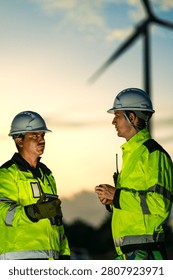 In dim orange afterglow, two technicians face to face stand talking with radios clipped on on open ground below the towers. Renewable electricity, green engineering careers and climate.
