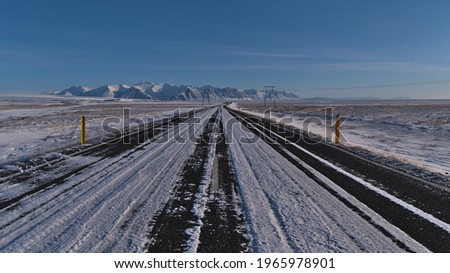 Diminishing perspective of icy paved ring road (route 1) in the south of Iceland near Jökulsárlón on sunny day in winter season with power pylons and rugged snow-capped mountains in background.