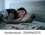 In a dimly lit bedroom, a woman lies awake in bed, scrolling through her smartphone. Her sleeplessness is evident as she struggles with nighttime phone dependency.