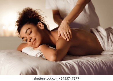 In a dimly lit spa, a black woman relishes the comfort of a therapeutic back massage performed with expertise - Powered by Shutterstock