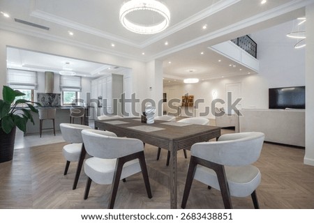 The dining area features a spacious table surrounded by white chairs, illuminated by modern lighting fixtures. Large windows enhance the bright atmosphere, showcasing a contemporary design