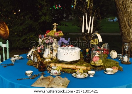 The dining table is decorated in the style of Alice in Wonderland. An open old book, a cake, tea bowls, candles in a candlestick, an hourglass on a blue tablecloth late in the evening.