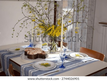 Dining table set for easter with traditional colomba pasquale, mimosa centerpiece, decorative eggs and white rabbit figurine, bright spring celebration ready scene