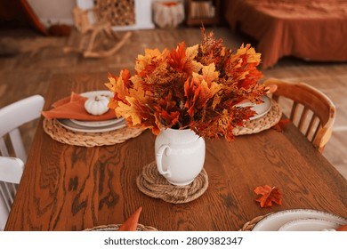 A dining table is set with a white vase filled with orange and yellow leaves. A small white pumpkin sits nearby. The room has wooden floors and a warm atmosphere.
