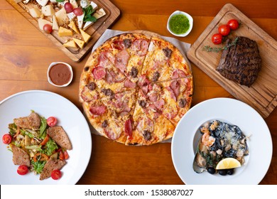 Dining Wooden Table With Food, Top View. Pizza, Salad, Cheese Board, Steak, Black Seafood Pasta. Concept For Restaurant Menu.