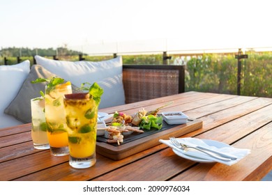 Dinner on a wooden table with sofa and pillow seat in an open air hotel restaurant with grass field background at sunset time with a clear sky, copy space for text.