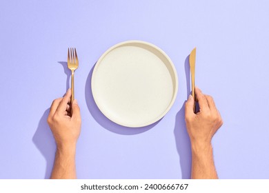 Dinner place setting. White empty plate and hand holding gold fork and knife on lilac background - Powered by Shutterstock