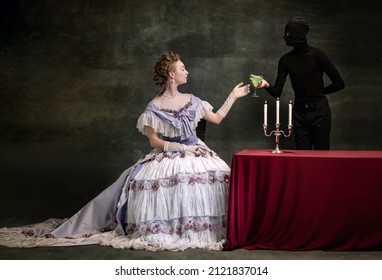 Dinner time. Cinematic portrait of young beautiful woman in image of medieval royal person in renaissance style dress and her servant, page isolated on dark vintage background. Comparison of eras