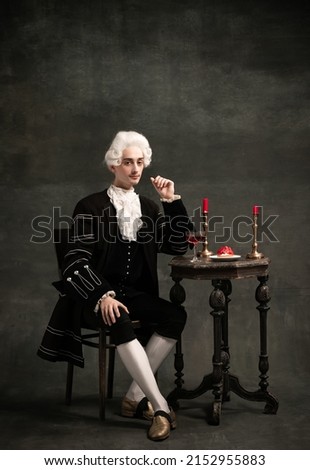 Dinner. Young man wearing wig and vintage medieval outfit like famous composer isolated on dark green vintage background. Retro style, fashion, art, comparison of eras concept.