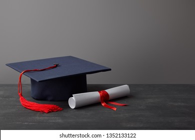 Diploma And Academic Cap With Red Tassel On Dark Surface On Grey