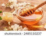 Dipping wooden spoon into glass bowl with honey, acacia flowers are on wooden table near. Top view.