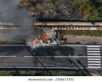 A direct drone view of a massive fire raging on the side of a road. Flames and thick smoke rise from burning debris near large industrial chimneys. The asphalt is covered in soot stains.