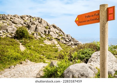 Directional signpost towards sun-drenched Fóia mountaintop, showcasing hiking trails PR 3 Trilho da Fóia and PR 5 Percurso das Cascatas, with a misty cityscape of Portimão in the far-off distance. - Powered by Shutterstock