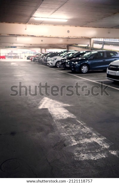 Directional white arrow on a floor of a parking lot\
and cars out of focus in the background. Selective focus. Busy car\
park of a shopping mall. Parking cost and vehicle density problem\
in town.
