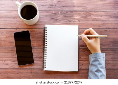 Directly Above Hand Holding Pen With Blank Note Pad, Smart Phone And Cup Of Coffee