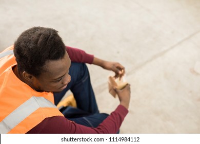 Directly Above Of Pensive Calm African-American Construction Worker In Orange Vest Sitting On Ground And Eating From Box During Lunch Break