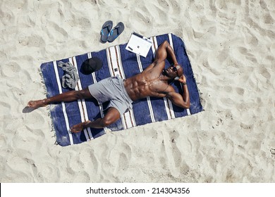 Directly above shot of  muscular young african guy sunbathing on beach. Man wearing sunglasses listening to music on while lying on a beach mat outdoors.