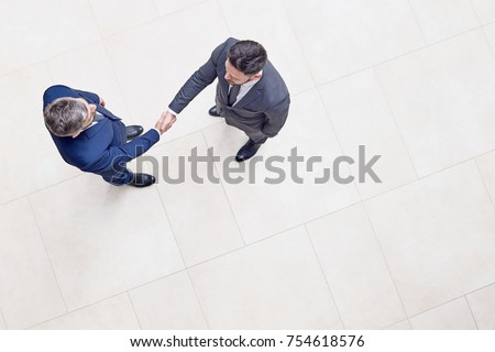 Directly above view of confident business partners in classical suits shaking hands after successful completion of negotiations, interior of office lobby on background