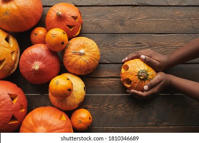 Directly From Above View Flat Lay Shot Of Carved Pumpkins And Tangerines With Jack O' Lantern Faces And Womans Hands Holding One Pumpkin For Halloween On Dark Wooden Table Background...