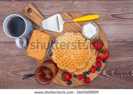 Directly above view of stack of toasted, heart-shaped Norwegian waffles, surrounded by fresh block of caramelized, brown cheese (brunost), cream and fresh berries on a wooden cutting board with coffee