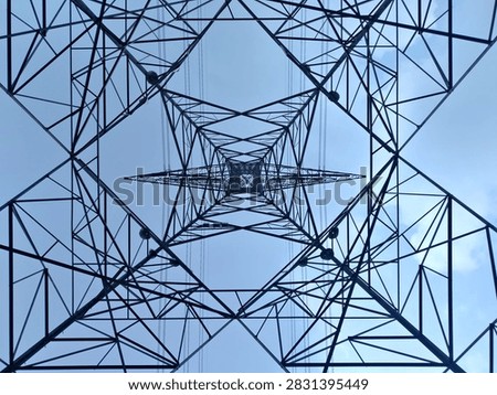 Directly looking up from inside a high voltage steel transmission tower pylon, showcasing symmetrical geometric lattice framework, steel beams, and power lines against a clear sky. Industrial infrastructure for electricity supply and energy grid.