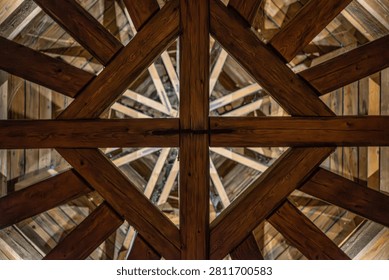 A directly upward, low-angle view of a complex wooden roof structure, featuring dark brown intersecting beams that form intricate geometric patterns in the ceiling of a tower or pavilion.