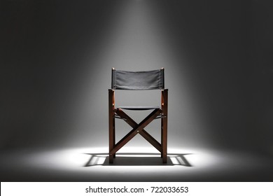 A Directors Chair Sits Against A Plain Dark Background Illuminated By A Single Overhead Spotlight.