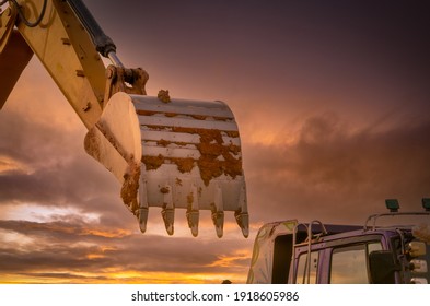 Dirt Metal Bucket Of Backhoe After Digging Soil. Backhoe Parked Near Truck With Golden Sunset Sky Background. Crawler Excavator. Earthmoving Machine At Construction Site At Dusk. Excavation Vehicle.