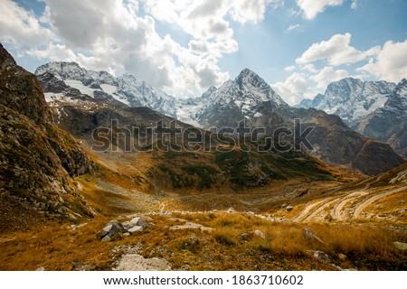 Dirt mountain road, serpentine with a cliff and a beautiful panoramic view of the gorge. Snow-capped mountain peaks, peaks cutting through the blue sky with clouds. The yellow autumn forest conveys th