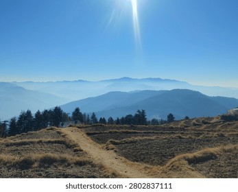 A dirt path cutting through hilly terraced fields with misty blue mountain layers beneath a bright sun.