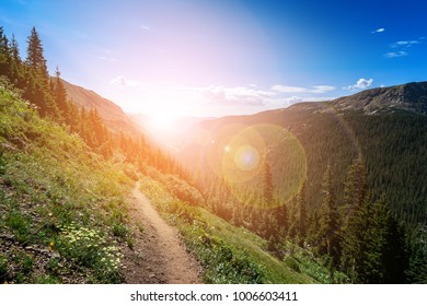 Dirt path hiking trail climbs through the Colorado mountains with the colorful light of the bright sun shining over the distant horizon