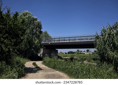 Dirt  path next to a brook bordered by trees and reeds with a viaduct passing over it in the italian countryside
