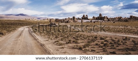 A dirt road cuts through a sparse, arid landscape near Potosi, Bolivia. Unique rock formations punctuate the terrain, with mountains visible in the distance under partly cloudy sky. Copy ad text space