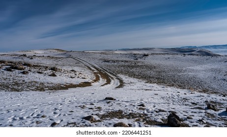 A Dirt Road Runs Along A Snow-covered High-altitude Plateau. Tire Tracks Bend And Go To Infinity. There Are Stones Scattered Around. A Mountain Range In The Distance. Blue Sky With Radial Clouds. Alta