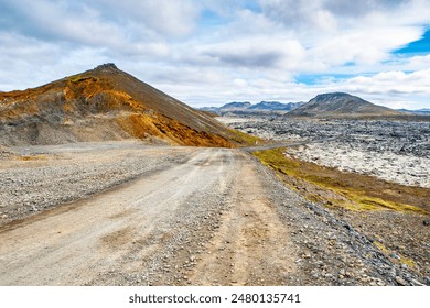 A dirt road winds through the stark and rugged landscape of solidified lava fields under a cloudy sky on Icelands Reykjanes Peninsula, showcasing striking contrasts in the terrains color and texture. - Powered by Shutterstock