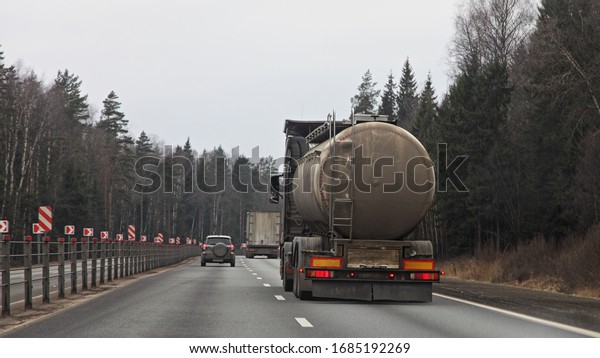 Dirty barrel semi truck rear view on\
suburban two-lane highway road at Spring day on forest background,\
liquid cargo\
transportation