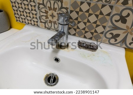 Dirty bathroom sink and mixer tap with soap scum and mold and featuring a filthy nail brush, a vintage tiled splashback and yellow painted wall
