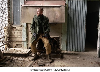 A Dirty Bearded Male Man Sits On An Engine Near The Entrance To An Abandoned Building Holding A Gun In His Hands. Doomsday Survivor Guarding The Entrance. Copy Space.