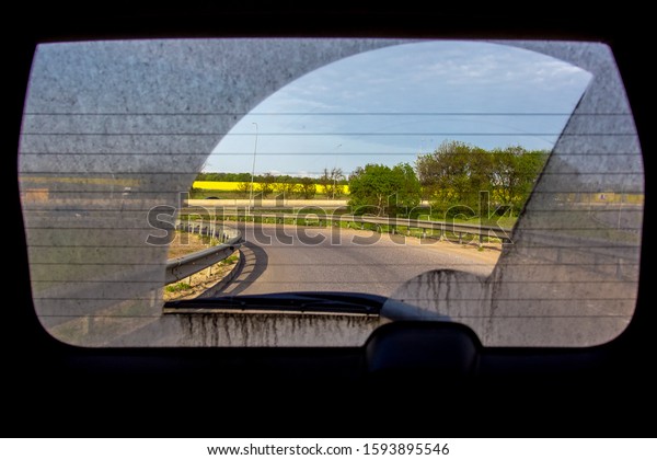 dirty car view from inside\
the car through the back window, rear window of a dirty car with a\
view of the asphalt road with turn road roadside with trees and\
field.