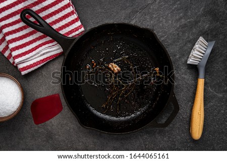 Dirty cast iron skillet being prepared for cleaning with coarse salt, brush, scraper and dish towel on a counter.
