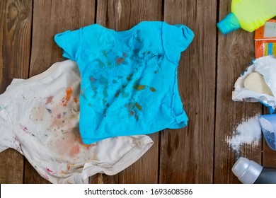 Dirty children's clothes are scattered on a wooden table next to washing powders and soap.Concept washing dirty spots, the best means of cleaning kids clothes. - Shutterstock ID 1693608586