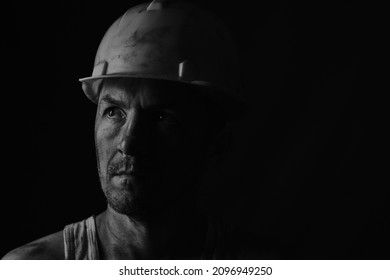 Dirty face of coal miner on a black background. Head of tired mine worker in a hard hat. Black and white photographic portrait