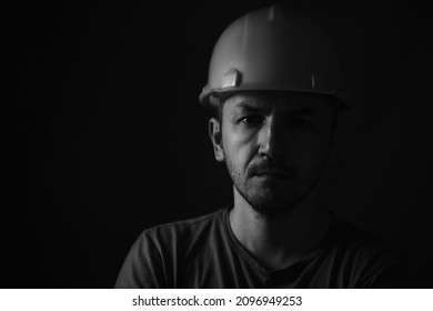 Dirty Face Of Coal Miner On A Black Background. Head Of Tired Mine Worker In A Hard Hat. Black And White Photographic Portrait