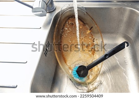 Dirty glass casserole with burnt-on fat is cleaned in the sink under a jet of water with a brush, dish washing by hand, tedious household chore, copy space, selected focus
