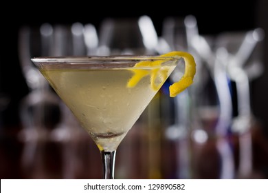 dirty martini chilled and served on a busy bar top with a shallow depth of field and color lights and glasses in the background garnished with a lemon twist
