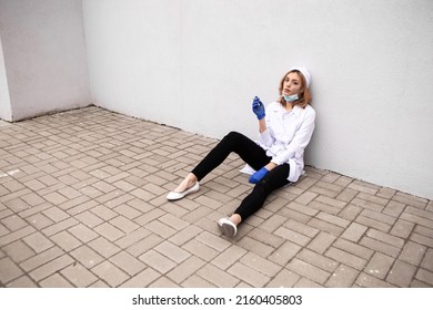 Dirty nurse with ash on face sitting outside hospital infirmary smoking cigarette after hard working day or surgery. Tired exhausted doctor woman dressed white medical gown have a rest due to stress