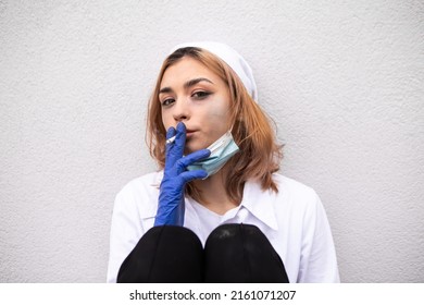 Dirty nurse with ash on face sitting outside hospital infirmary smoking cigarette after hard working day or surgery. Tired exhausted doctor woman dressed white medical gown have a rest due to stress

