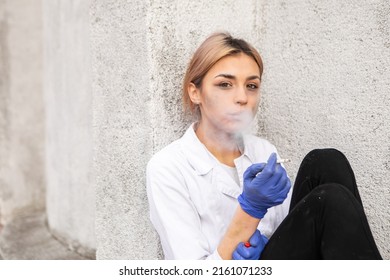 Dirty nurse with ash on face sitting outside hospital infirmary smoking cigarette after hard working day or surgery. Doctor woman dressed white medical gown, rubber blue gloves have a rest due to stre